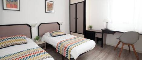 A hotel room with two beds, a desk, a chair, and a window with sheer curtains.