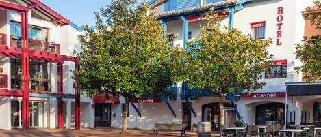 Un hotel con fachada roja y blanca, zona de estar al aire libre y un árbol en la entrada.