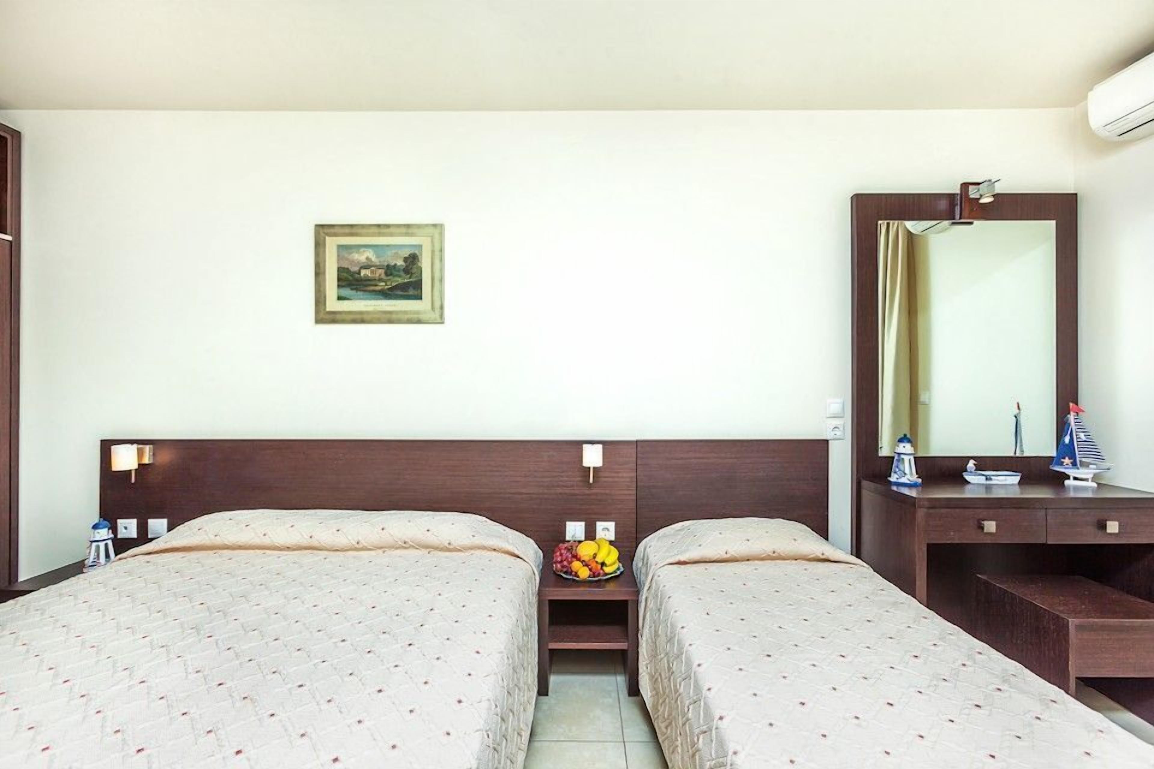 A hotel room with two beds, a small table with fruit, a painting on the wall, and a mirror above the dresser.