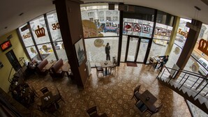 A spacious café with a high ceiling and large windows, featuring a herringbone floor pattern and a central seating area with tables and chairs. - Hotel Cisar (Lviv)