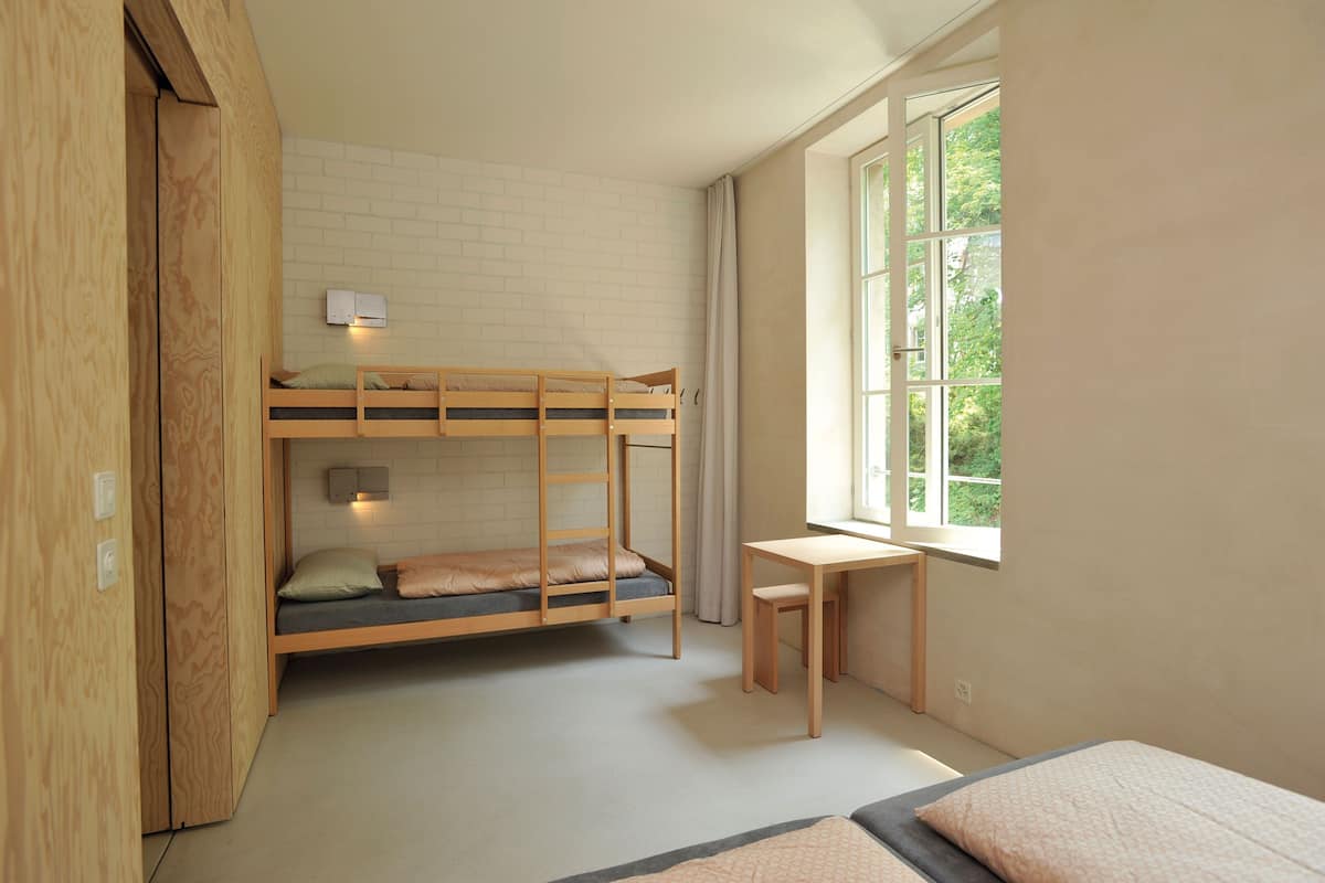 A compact room with a bunk bed, a small desk, and a window providing a view of greenery.