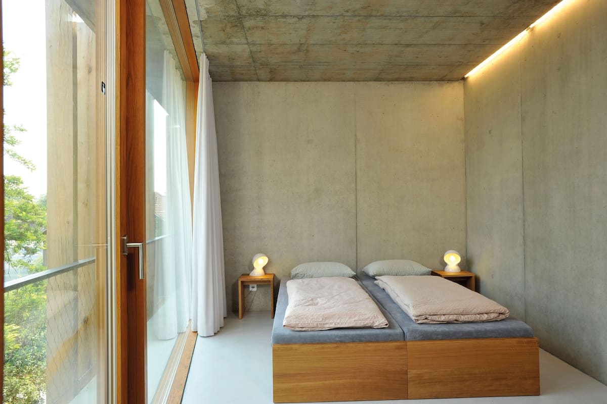 A bedroom with a wooden bed, two bedside tables with lamps, a window with curtains, and a concrete wall.