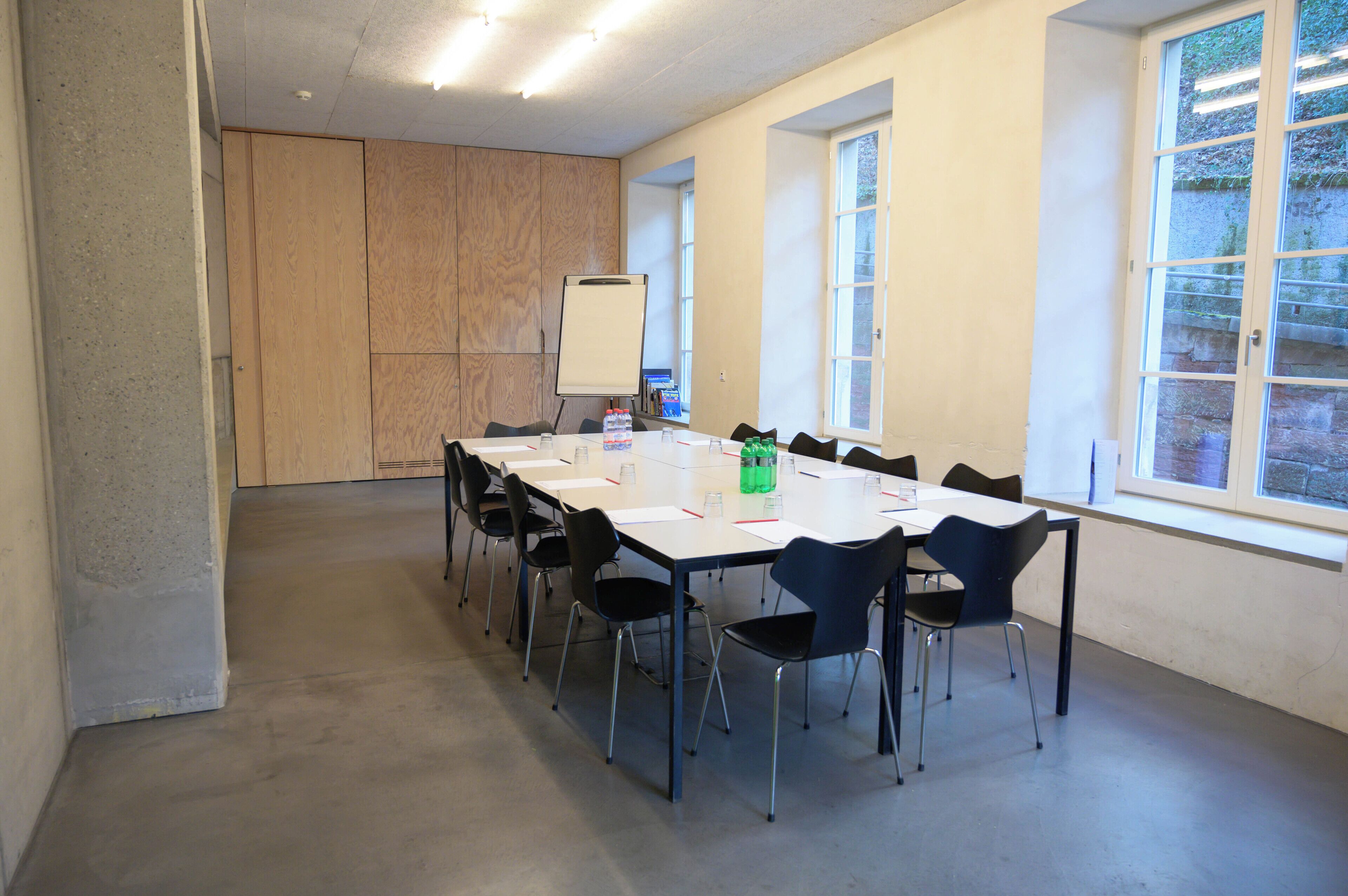 Uma sala de conferências com uma mesa comprida, cadeiras pretas, um quadro branco e janelas amplas.