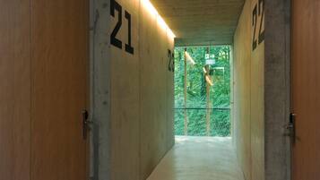 A corridor with wooden walls and numbered doors, leading to a glass door with a view of greenery outside.