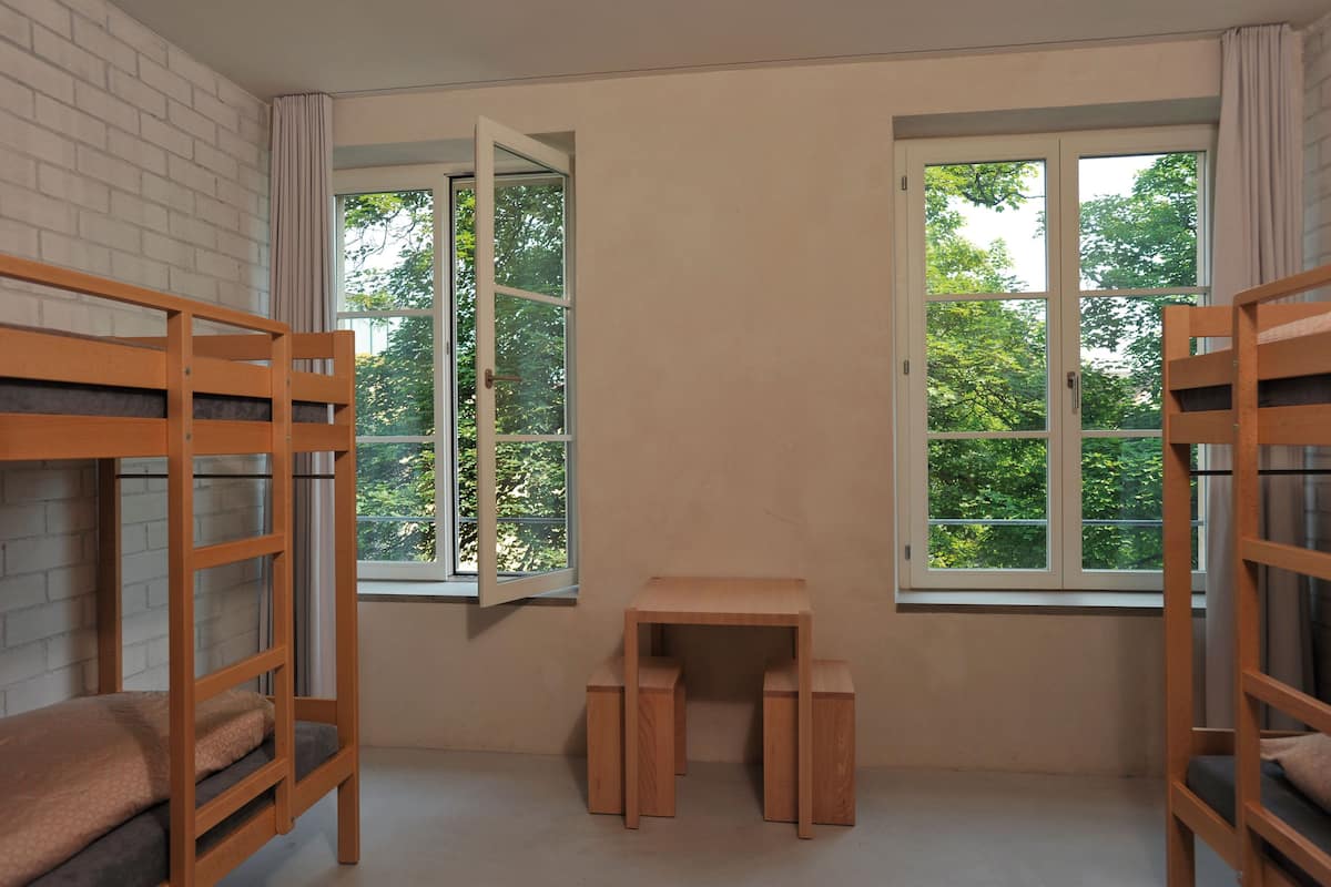 A room with bunk beds, a small wooden table, and two windows with curtains.