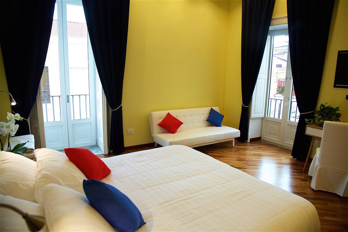 A hotel room with a large bed, a sofa, a chair, and a small table. There are curtains on the windows, and a plant is visible.