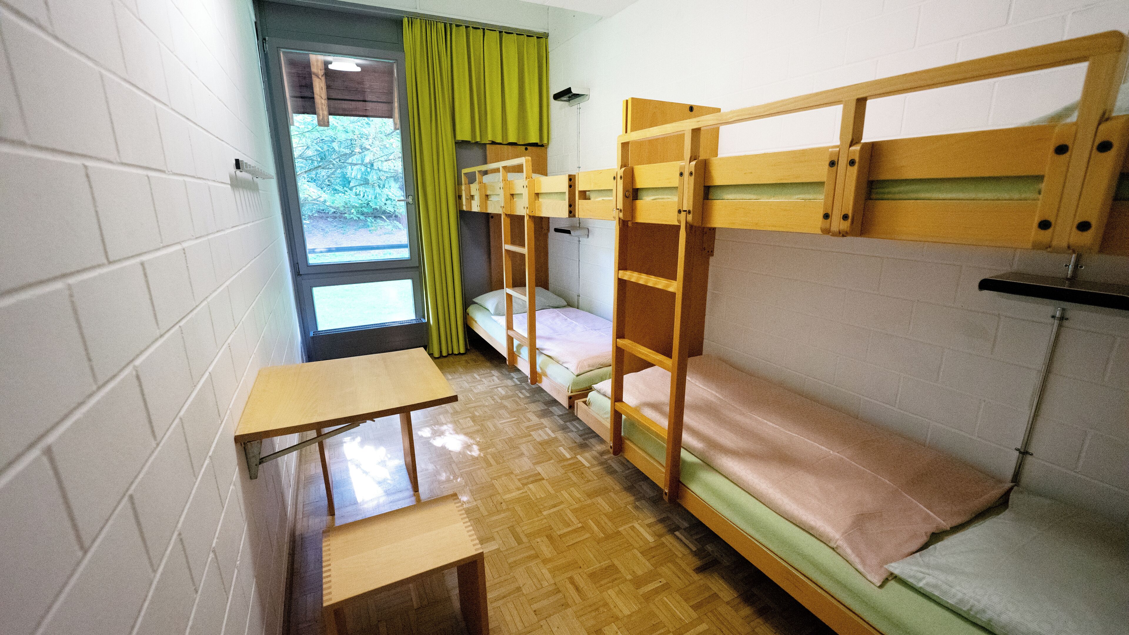 A dormitory room with bunk beds, a small wooden table, and a window with curtains.