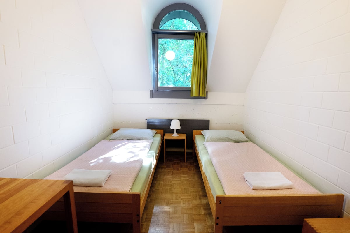 Una habitación pequeña y estrecha con dos camas individuales, una mesa de madera y una ventana que da a los árboles.