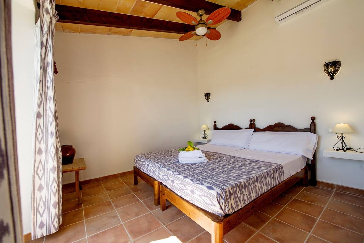 Une chambre avec un lit en bois, un ventilateur de plafond, une table de chevet avec une lampe et un luminaire fixé au mur.