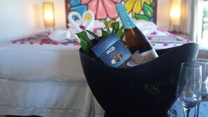 A bed with a floral headboard, a bedside table with a bottle and glasses, and a decorative flower arrangement. - Hotel Vila Jardim (Beberibe)