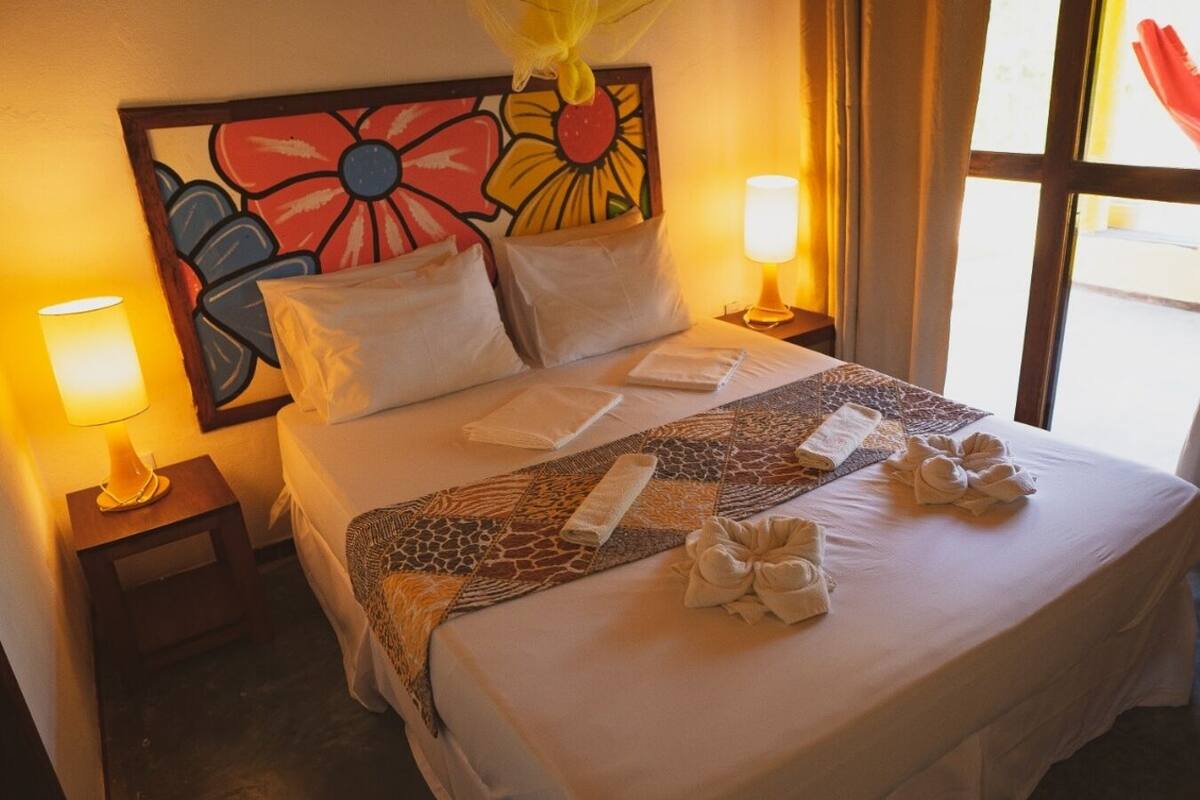Une chambre d’hôtel avec un lit, deux lampes de chevet, une tête de lit à motifs floraux et une fenêtre avec des rideaux.