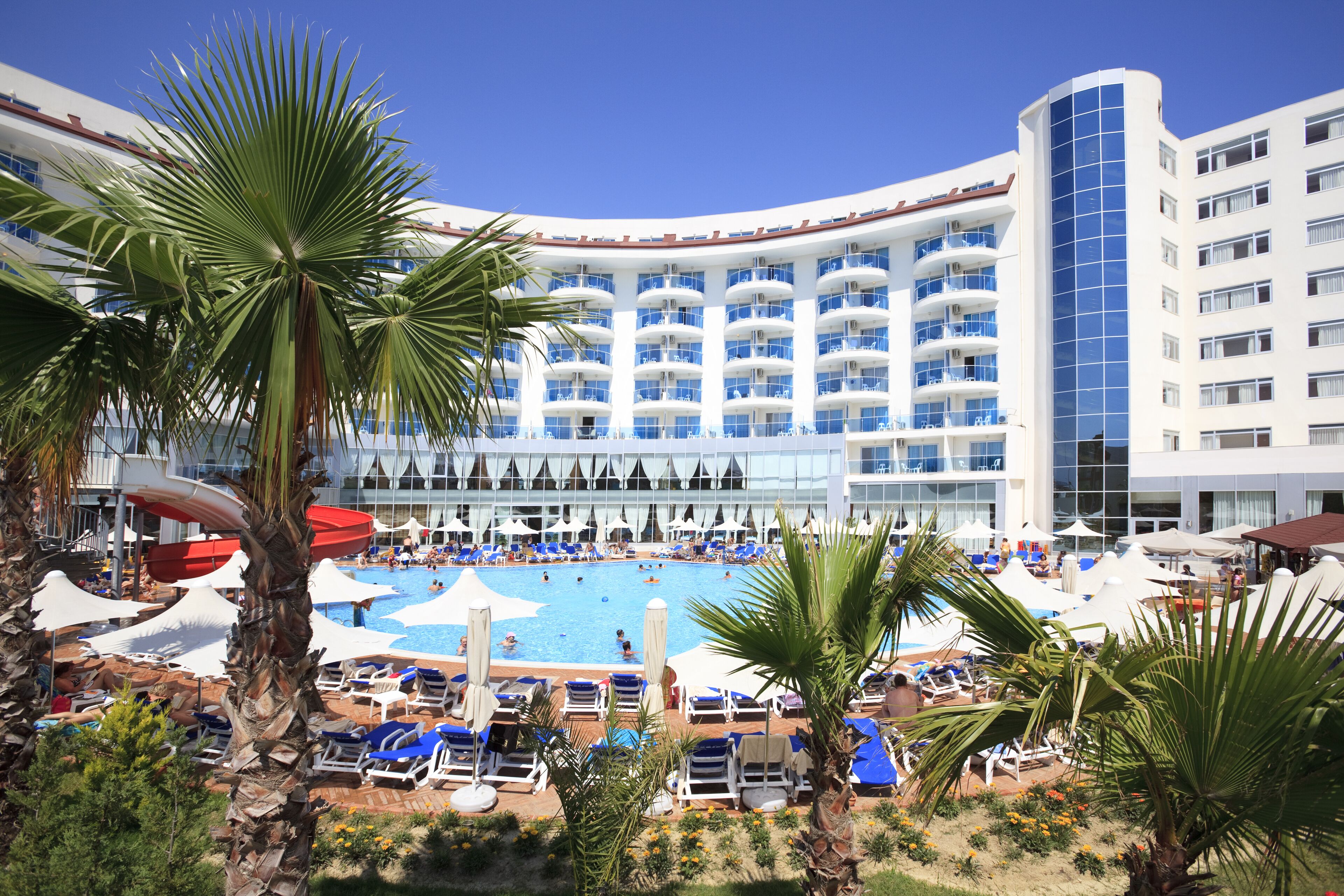 Um hotel com uma piscina grande, rodeada por espreguiçadeiras e guarda-sóis, palmeiras à beira da piscina e um edifício moderno com várias varandas.