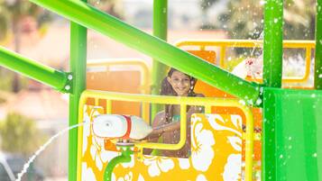 Un área de juegos con agua, con un niño jugando en un tobogán amarillo y naranja.