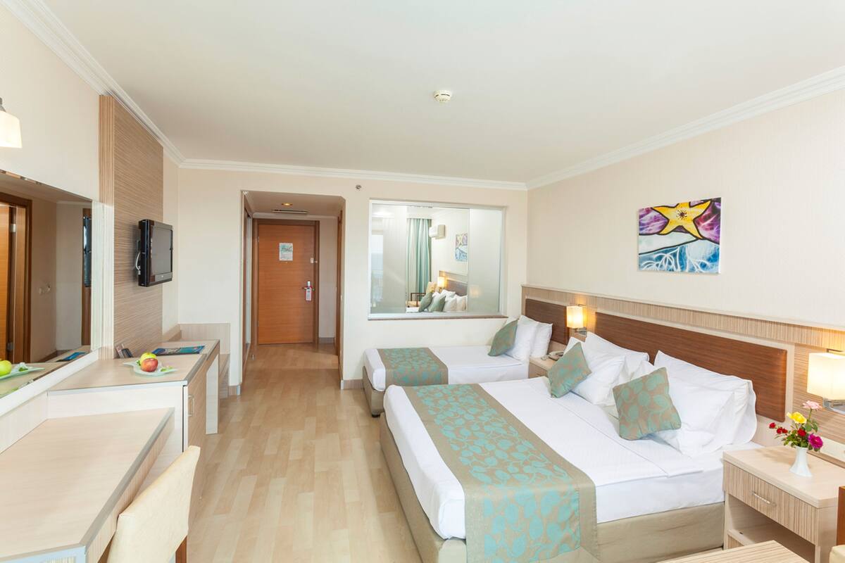 Habitación de hotel con dos camas, un escritorio, un televisor y un baño visible a través de una puerta de cristal.
