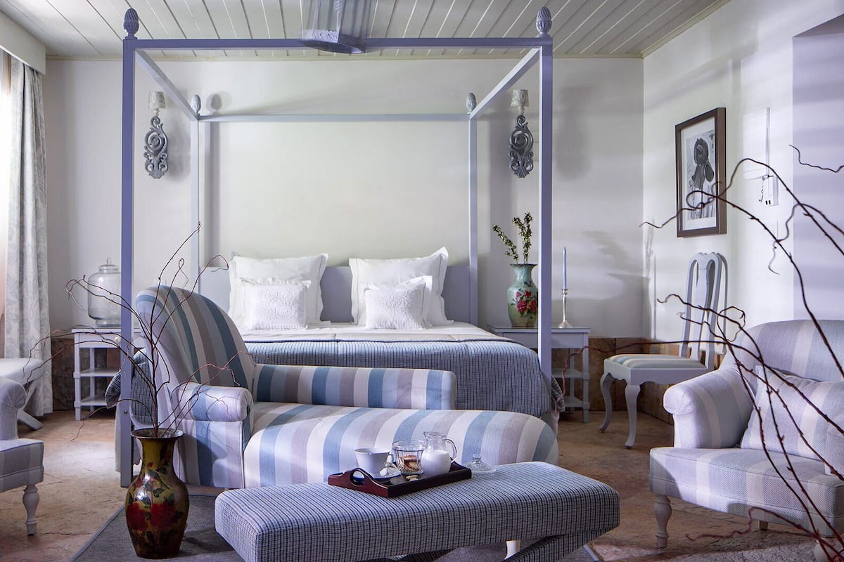 A four-poster canopy bed with white linens, striped armchairs, a small table with a vase, and a decorative vase on the floor.