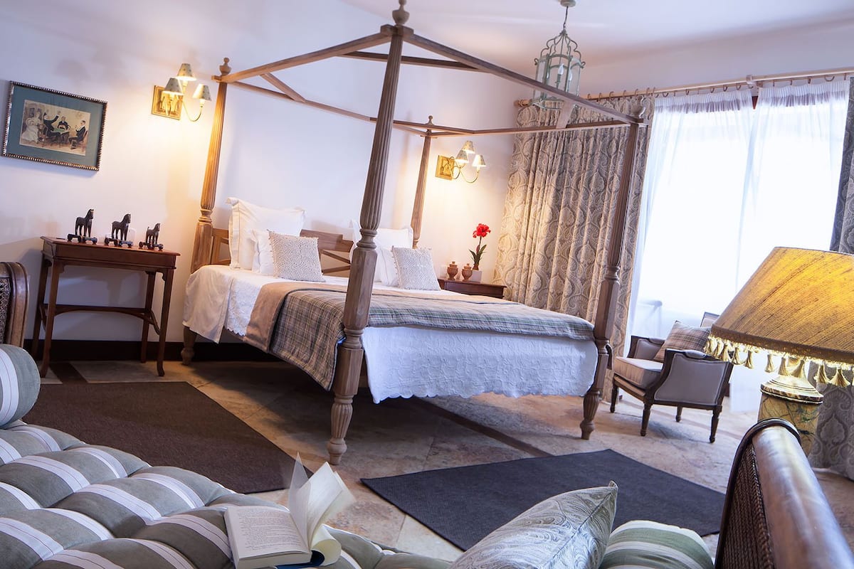 A four-poster bed with a canopy, a bedside table with a lamp, a chair, a sofa, and a framed picture on the wall.