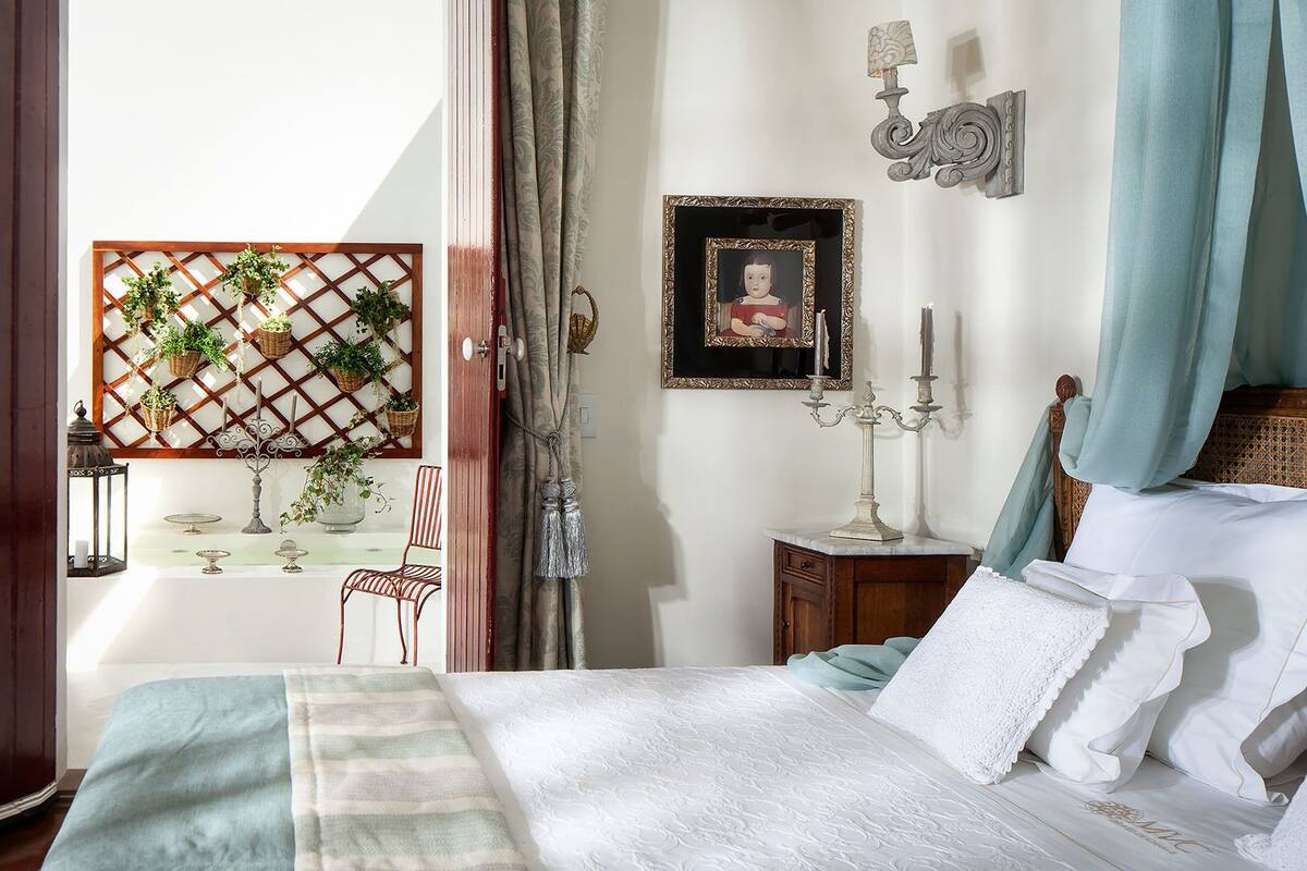 A bedroom with a bed, a nightstand, a wall-mounted lamp, a picture frame, and a window with curtains.