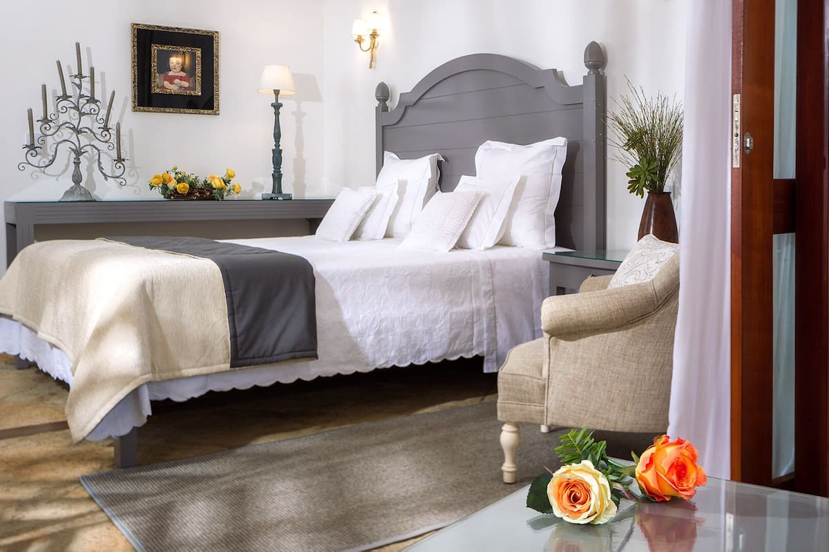 A neatly arranged bedroom with a bed, two chairs, a bedside table, a mirror, and a wall-mounted lamp.