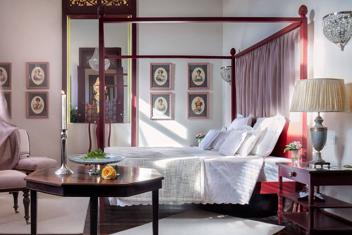 A four-poster bed with a white bedspread, a wooden nightstand with a lamp, a chair, and framed pictures on the walls.