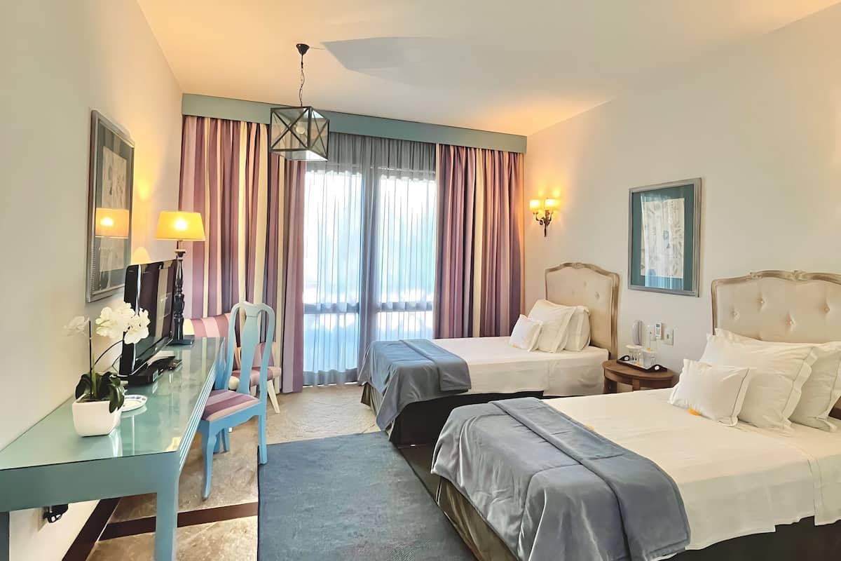 A hotel room with a glass desk, two beds, a chair, and a large window with curtains.