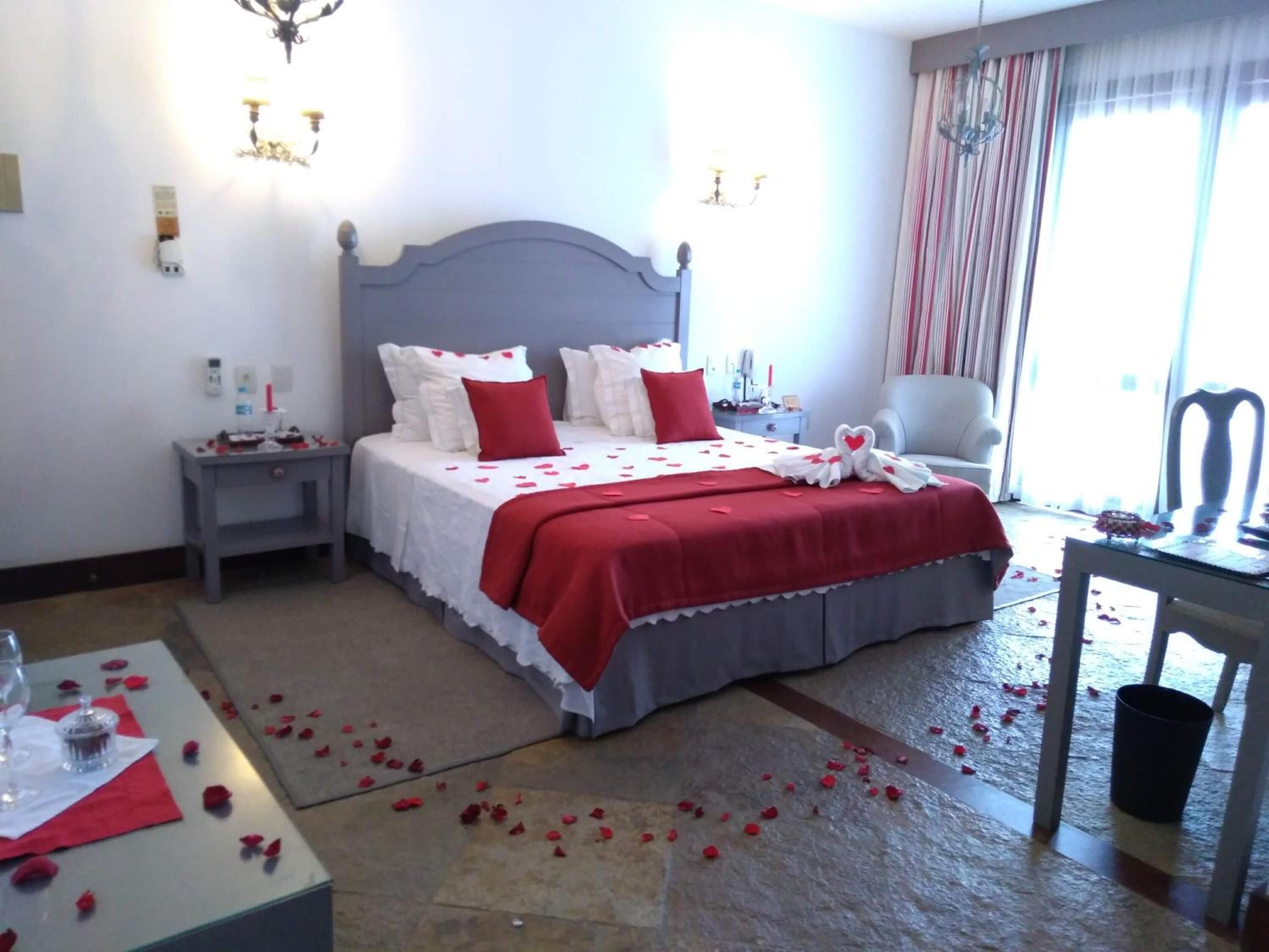 Quarto bem arrumado com cama, mesas de cabeceira, uma cadeira e uma pequena mesa com um copo.