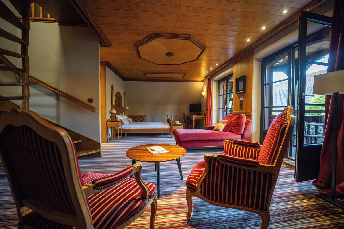 A hotel room with a wooden staircase, a bed, a red sofa, striped armchairs, and a small table.