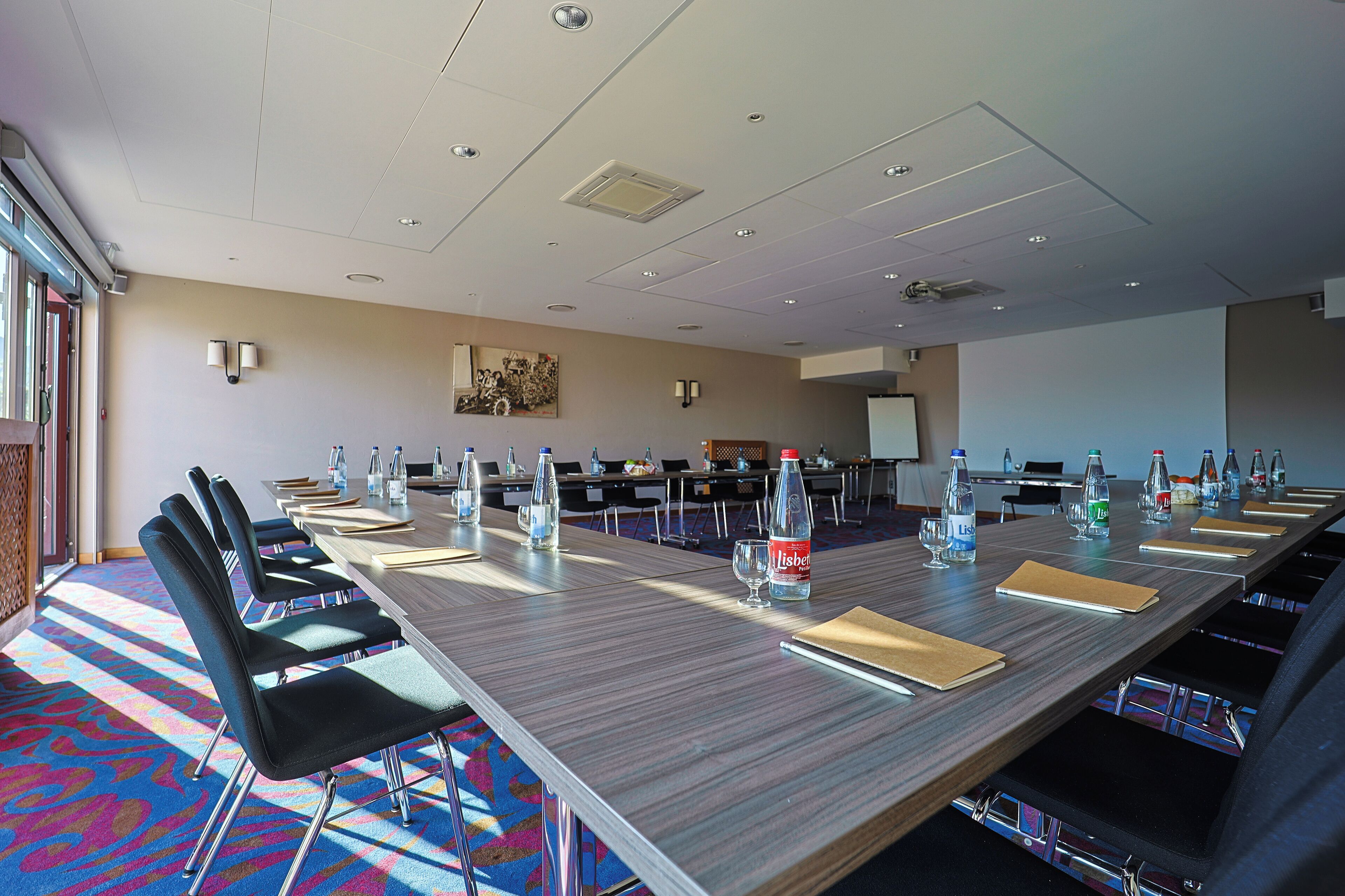 Ein Konferenzraum mit einem langen Tisch, Stühlen, Wasserflaschen und einer Projektionsleinwand.