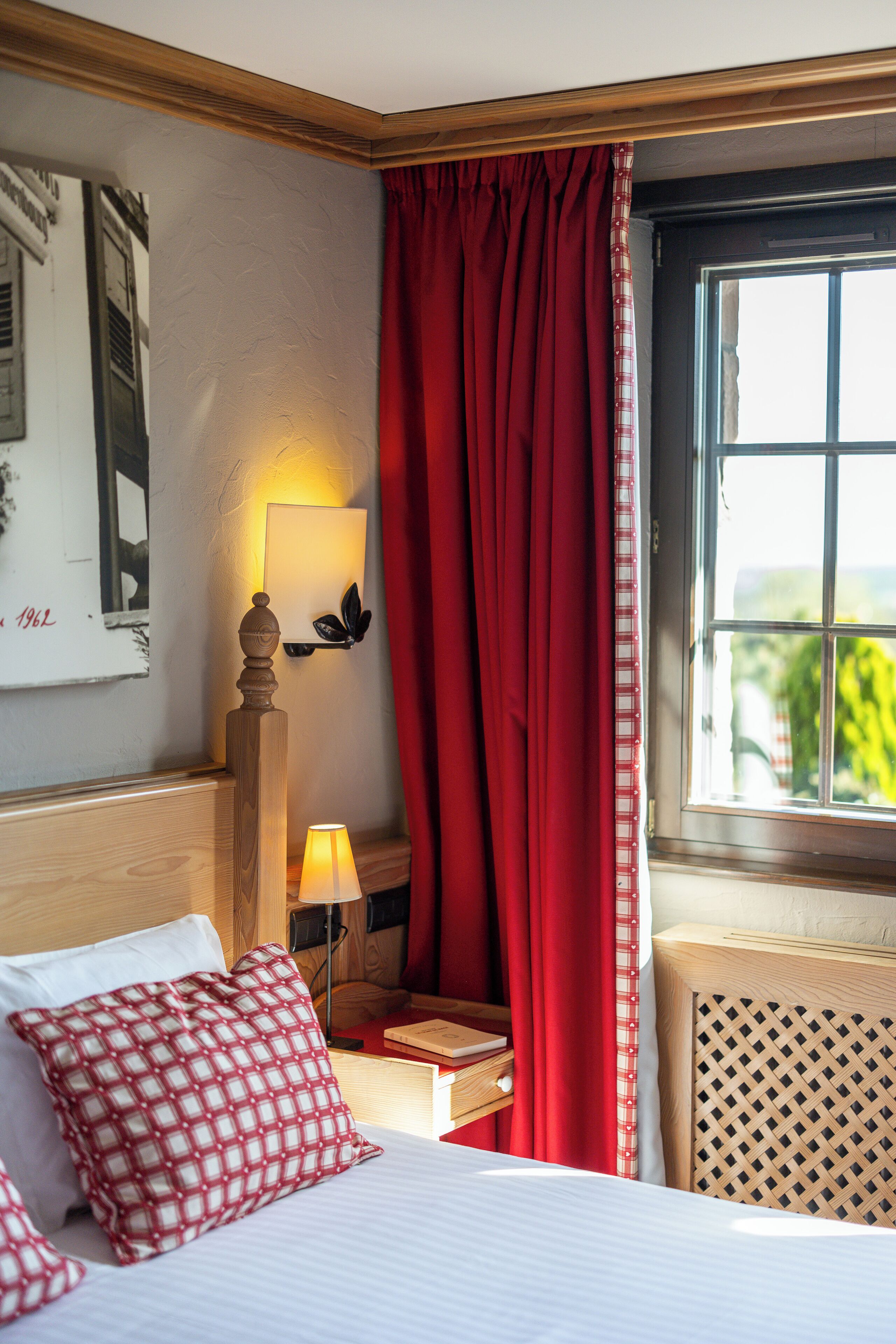 Hotellihuone, jossa on sänky, punaiset verhot, yöpöytä lampun kanssa ja ikkuna, josta on näkymä.