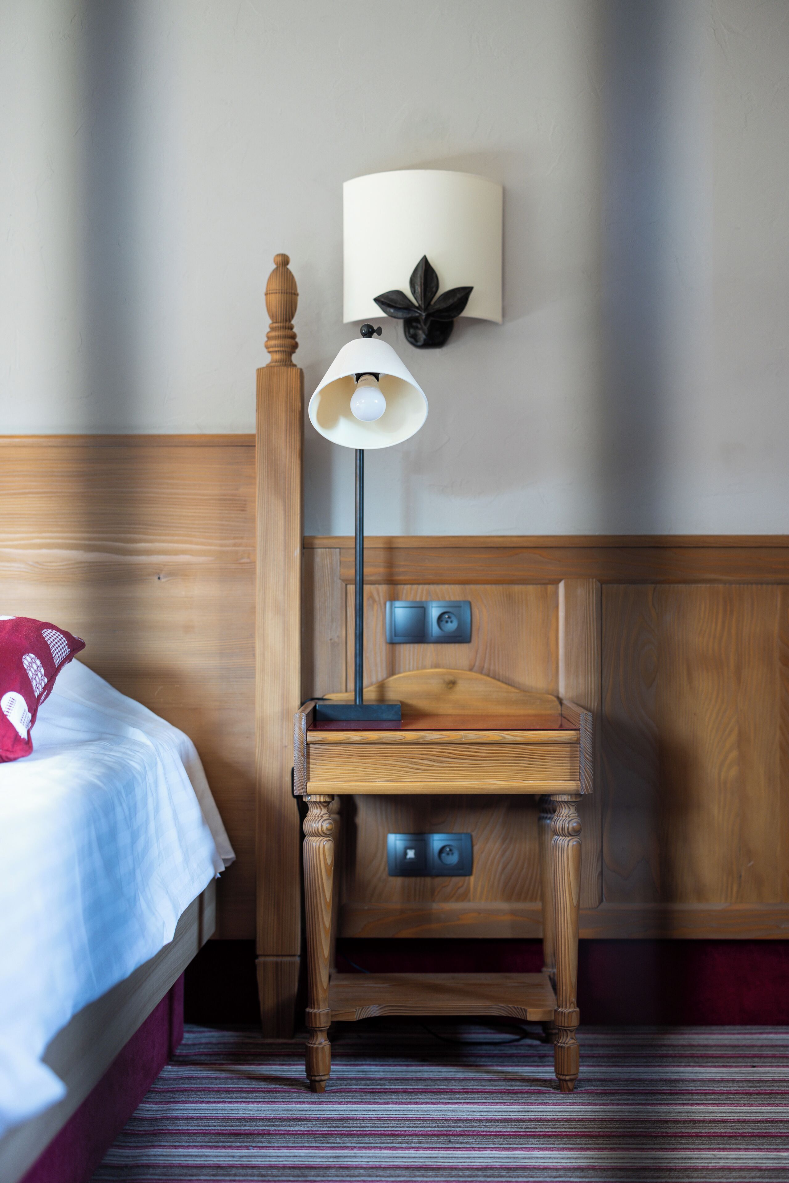 Une table de chevet en bois avec une lampe, un lit avec un oreiller rouge et un luminaire fixé au mur.