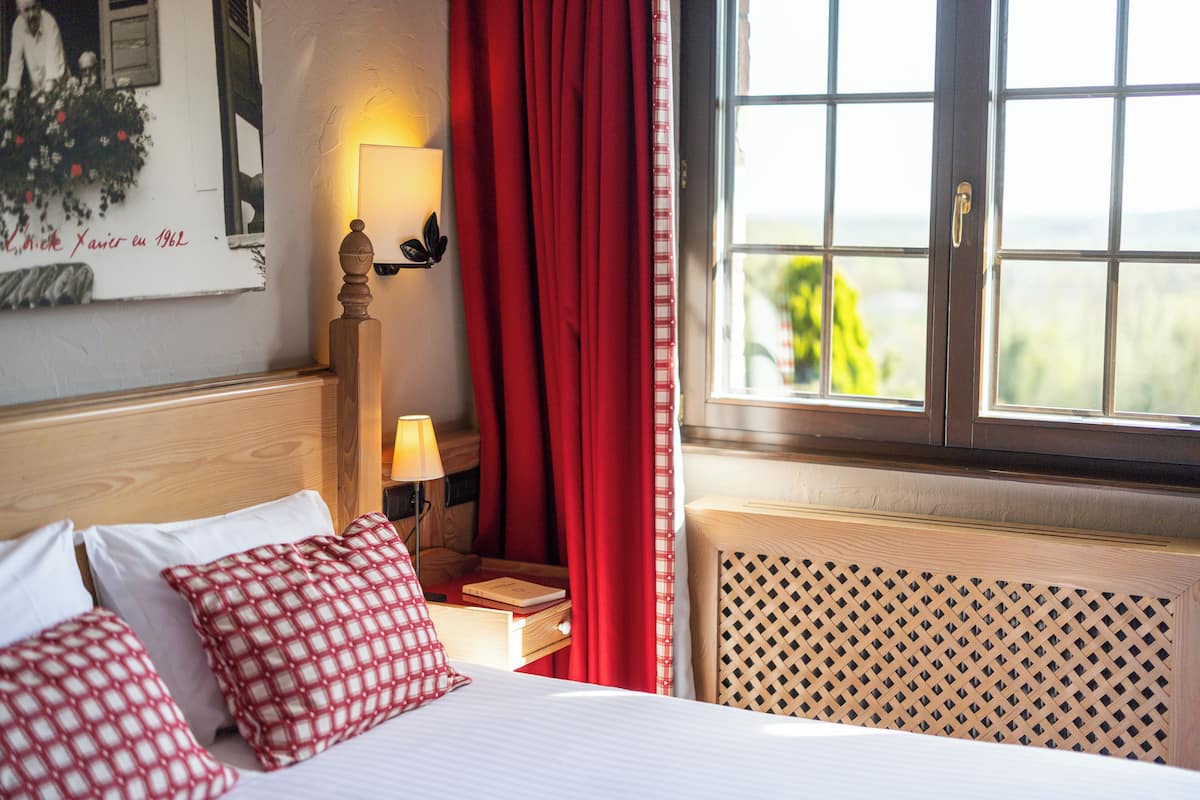 A hotel room with a bed, red curtains, a nightstand with a lamp, and a window with a view.