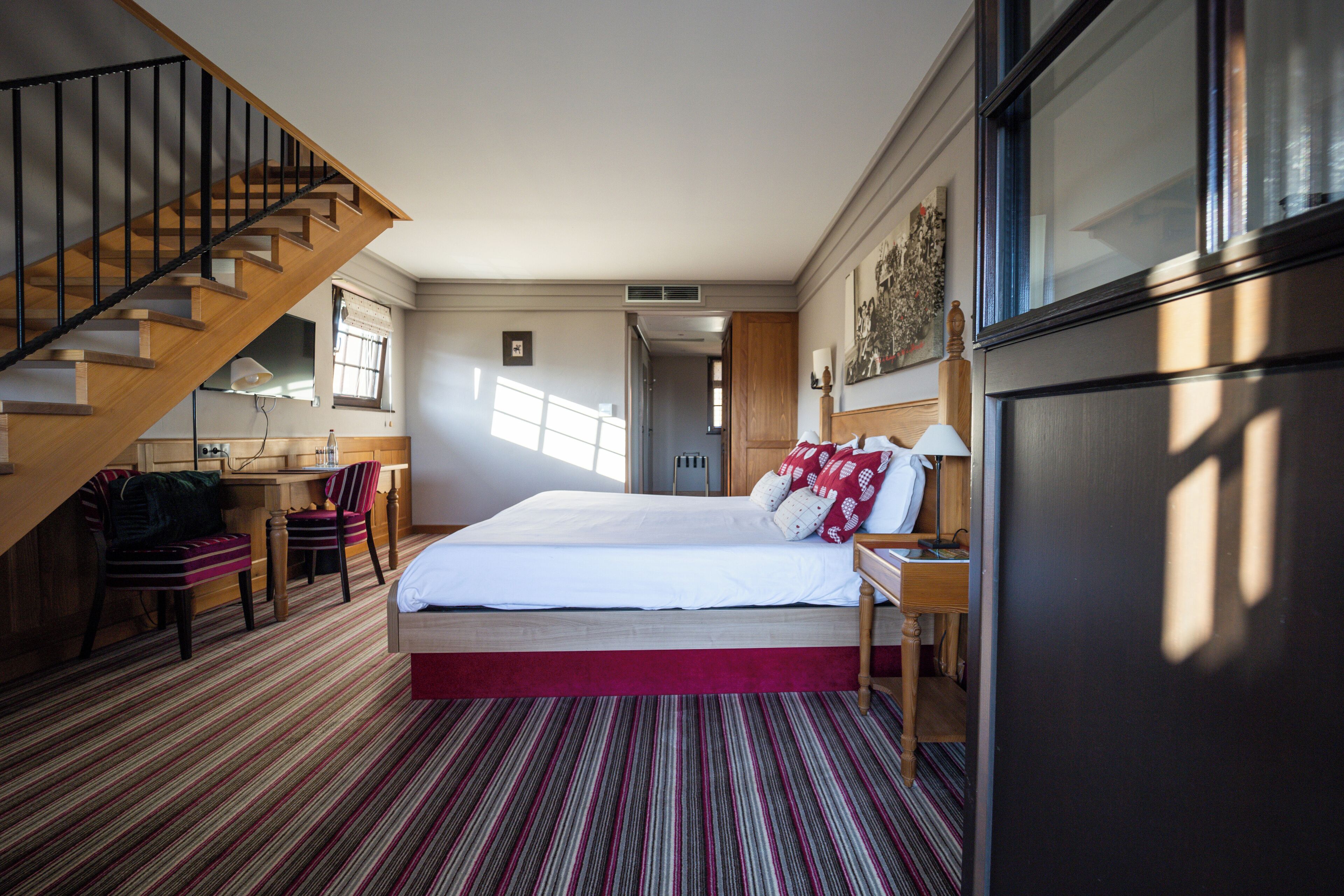 Ein Hotelzimmer mit einer Holztreppe, einem Bett mit rot-weißer Bettwäsche, einem Streifenmuster-Teppich, einem Schreibtisch mit Stuhl und einem großen Fenster.