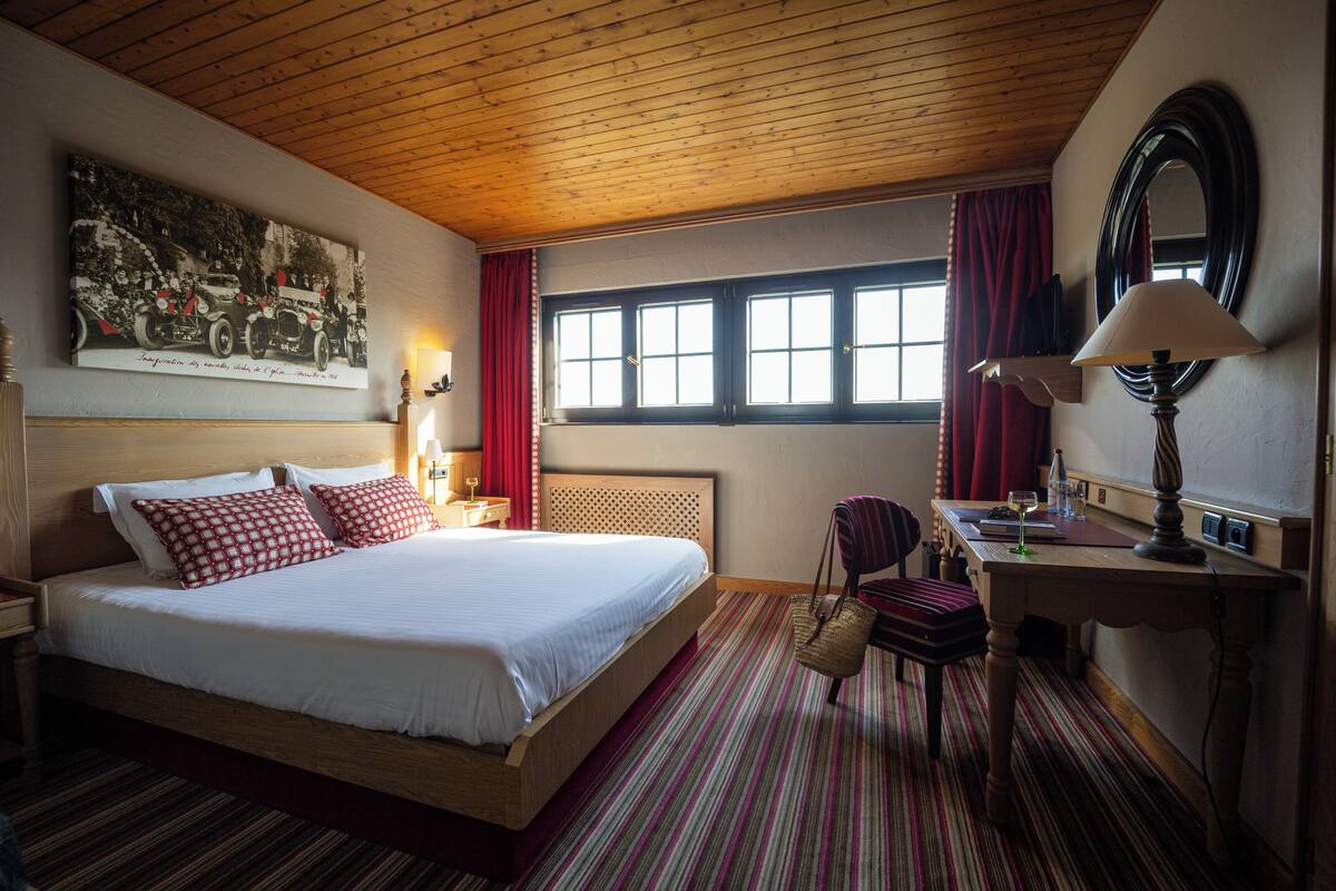 A hotel room with a wooden ceiling, a large bed with red and white bedding, a desk with a lamp, a chair, and a window with curtains.