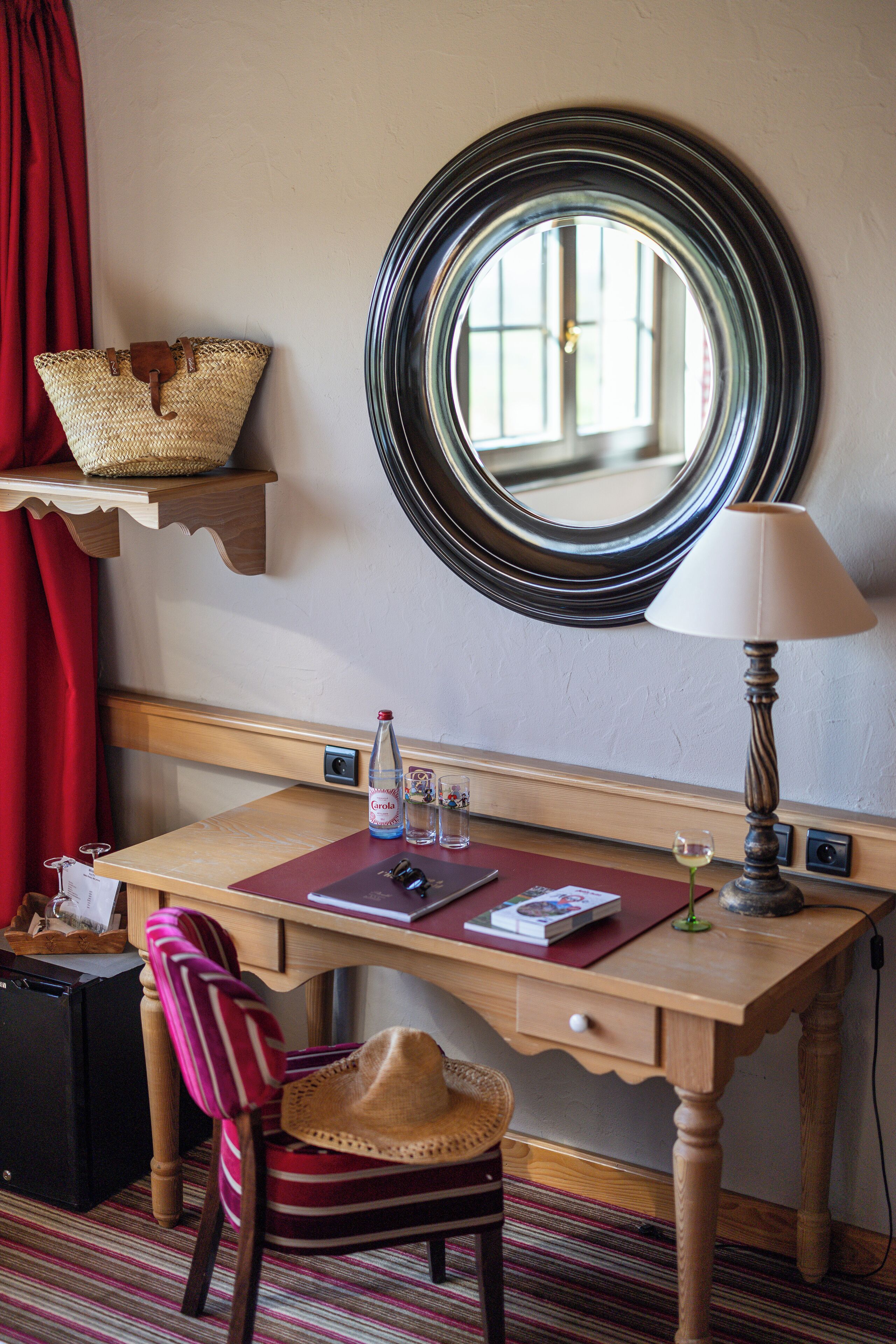 A wooden desk with a mirror, a lamp, a bottle of water, a glass, and a book. There is a chair with a hat and a red curtain in the background.