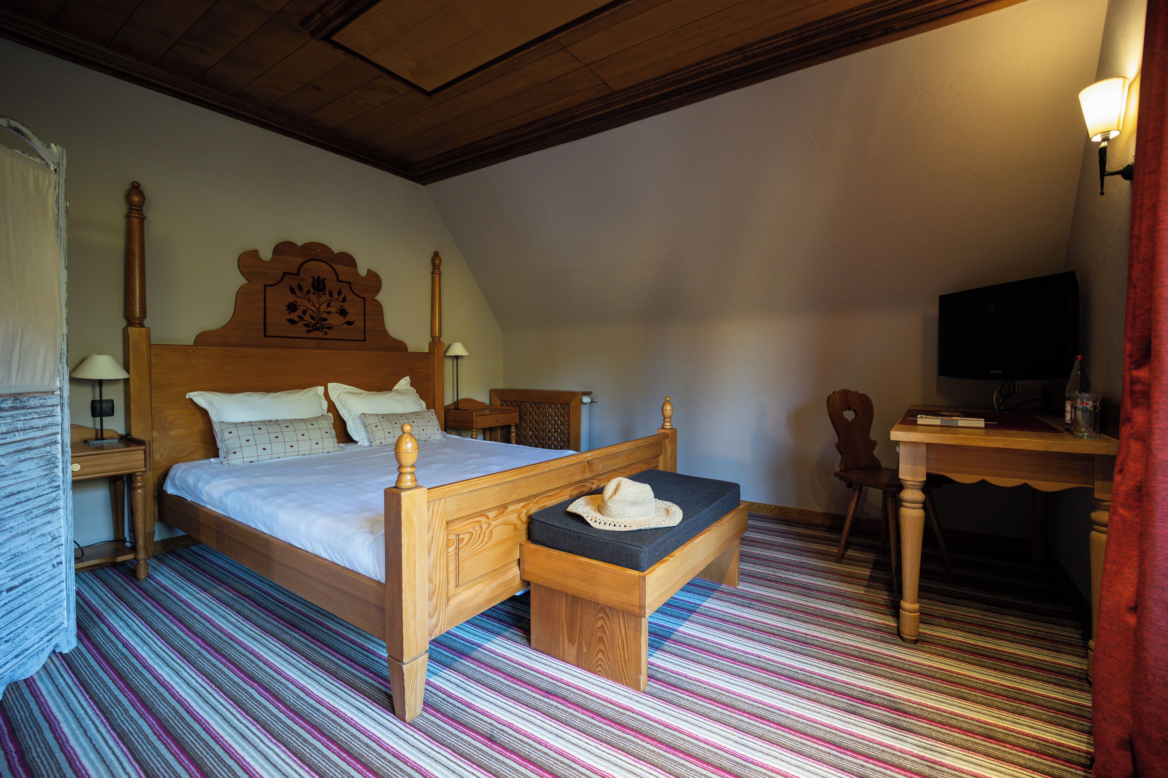 A bedroom with a wooden bed, a bench with a hat, a desk, and a television.