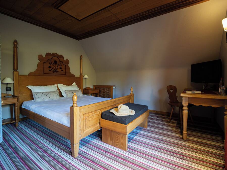 Ein Schlafzimmer mit einem hölzernen Bett, einer Bank mit einem Hut, einem Schreibtisch und einem Fernseher.