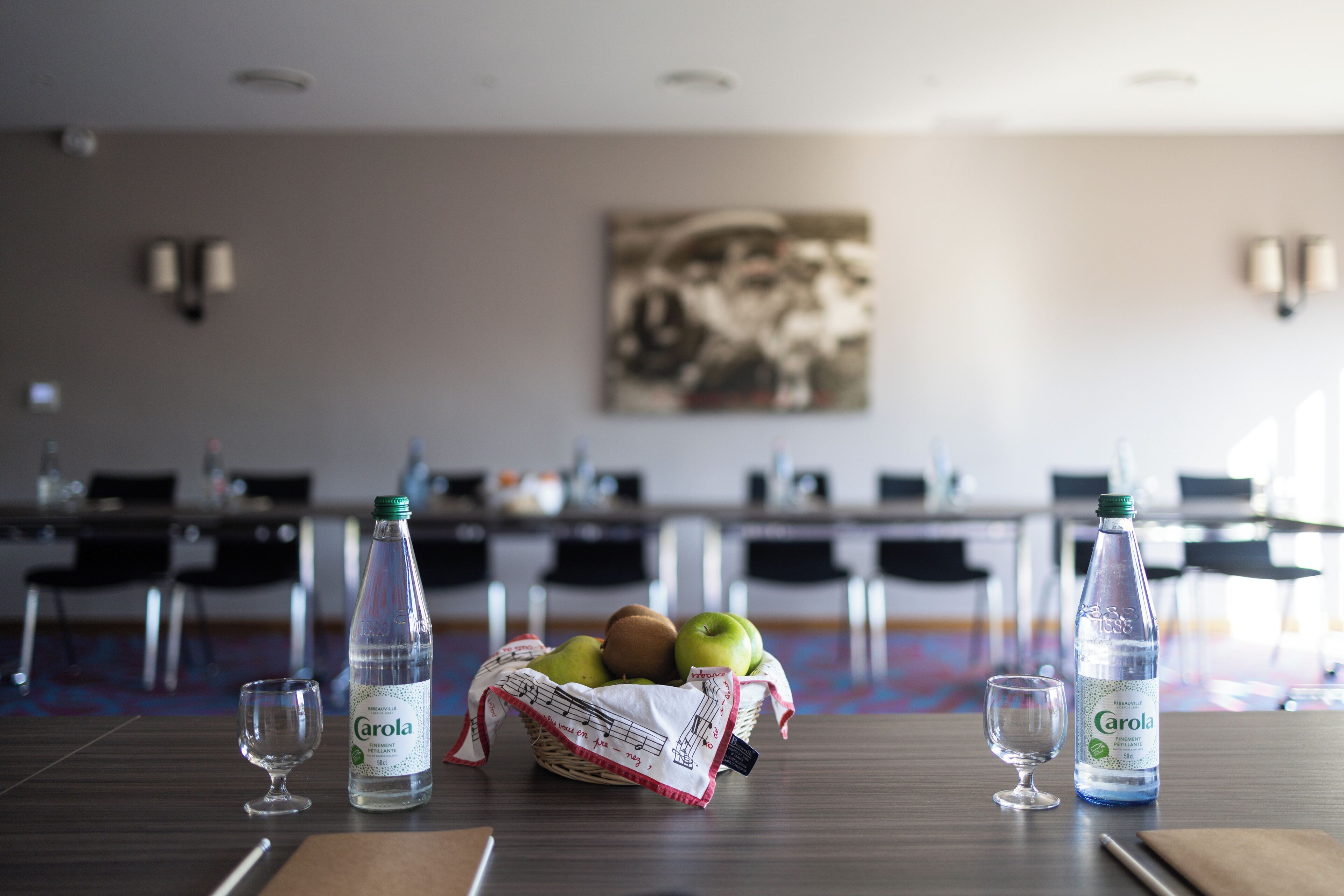 Une salle de conférence avec une table préparée pour une réunion, comprenant une bouteille d’eau Carola, un panier de fruits et deux verres à vin.