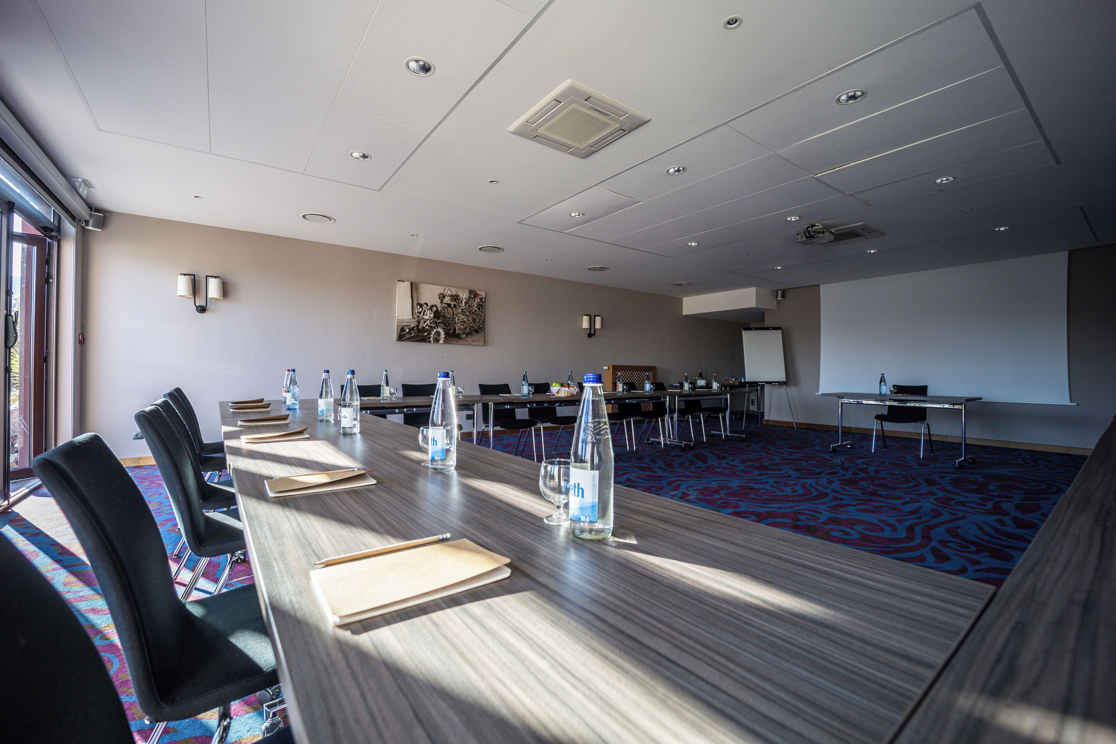 A conference room with a long table, chairs, water bottles, and a projector screen.