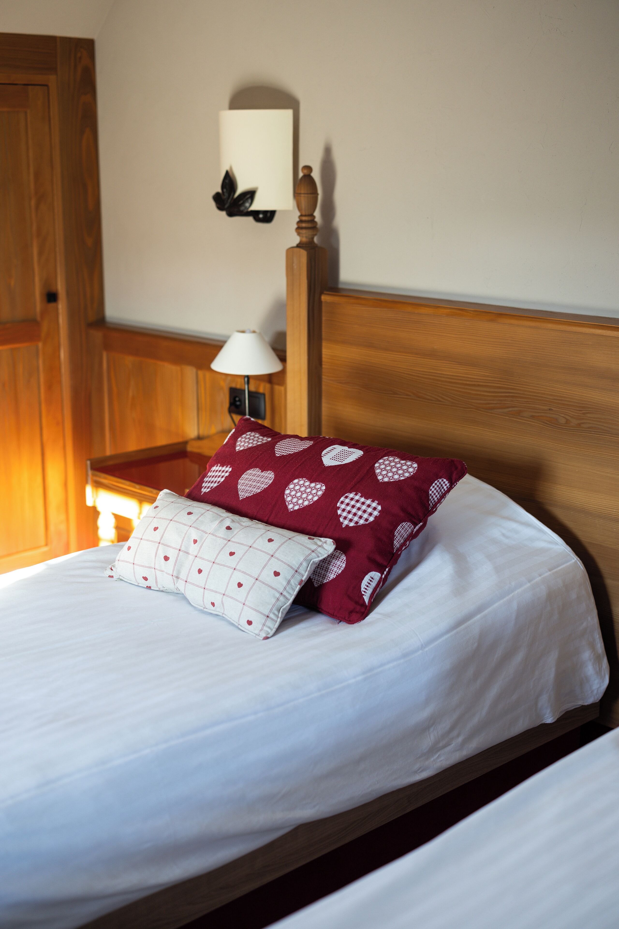 A neatly made bed with white bedding and two decorative pillows, a wooden headboard, a bedside table with a lamp, and a wall-mounted light fixture.