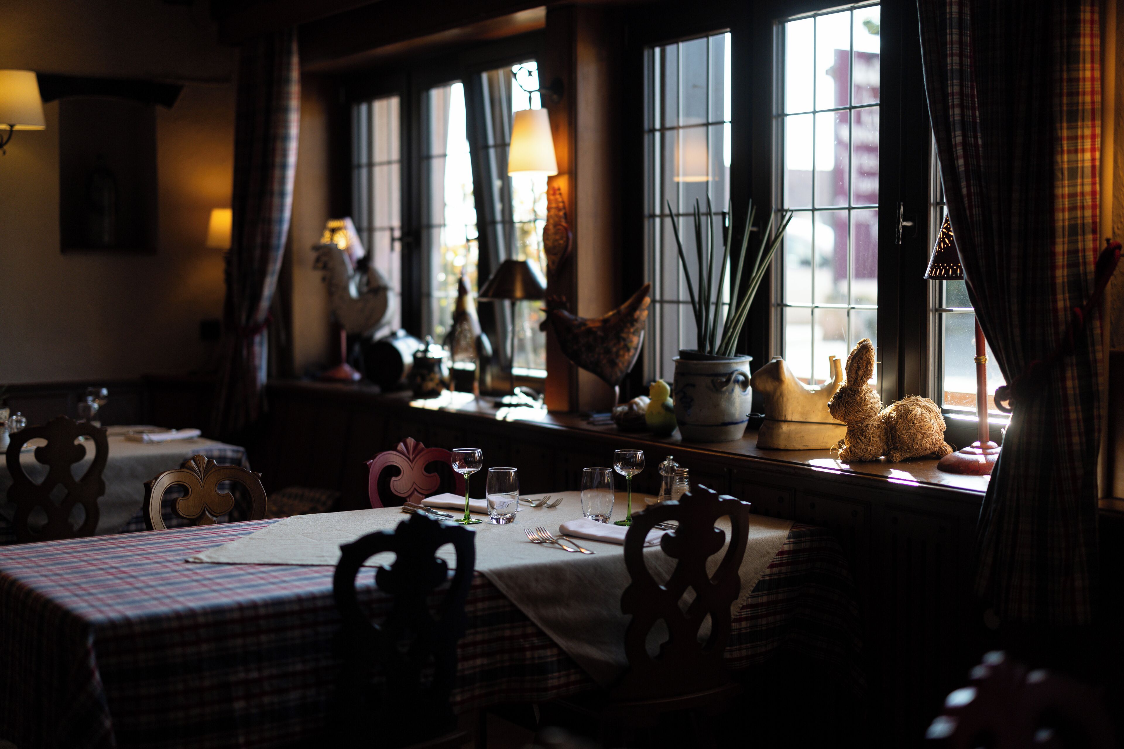 A dining area with a table set for two, featuring plaid tablecloths, wooden chairs, and a window with decorative items on the sill.