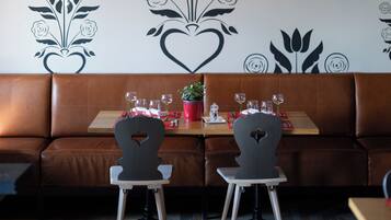 Een restaurantinterieur met een houten plafond, een muur met bloemen- en hartmotieven, een bruine leren zitbank en twee houten stoelen met een tafel ertussen.