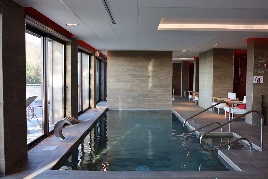 A modern indoor swimming pool with clear glass walls, a tiled floor, and a seating area with white chairs and red cushions.