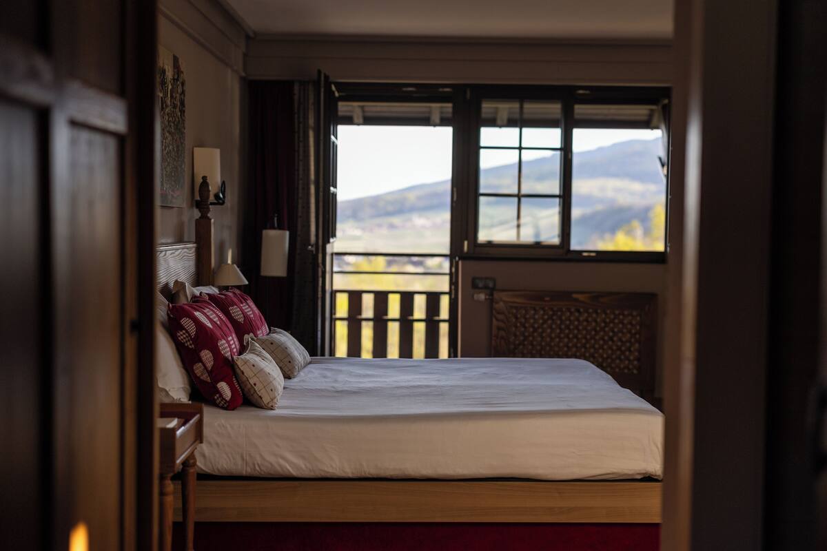 A bedroom with a bed, a nightstand, a lamp, a window with a view of mountains, and a door.