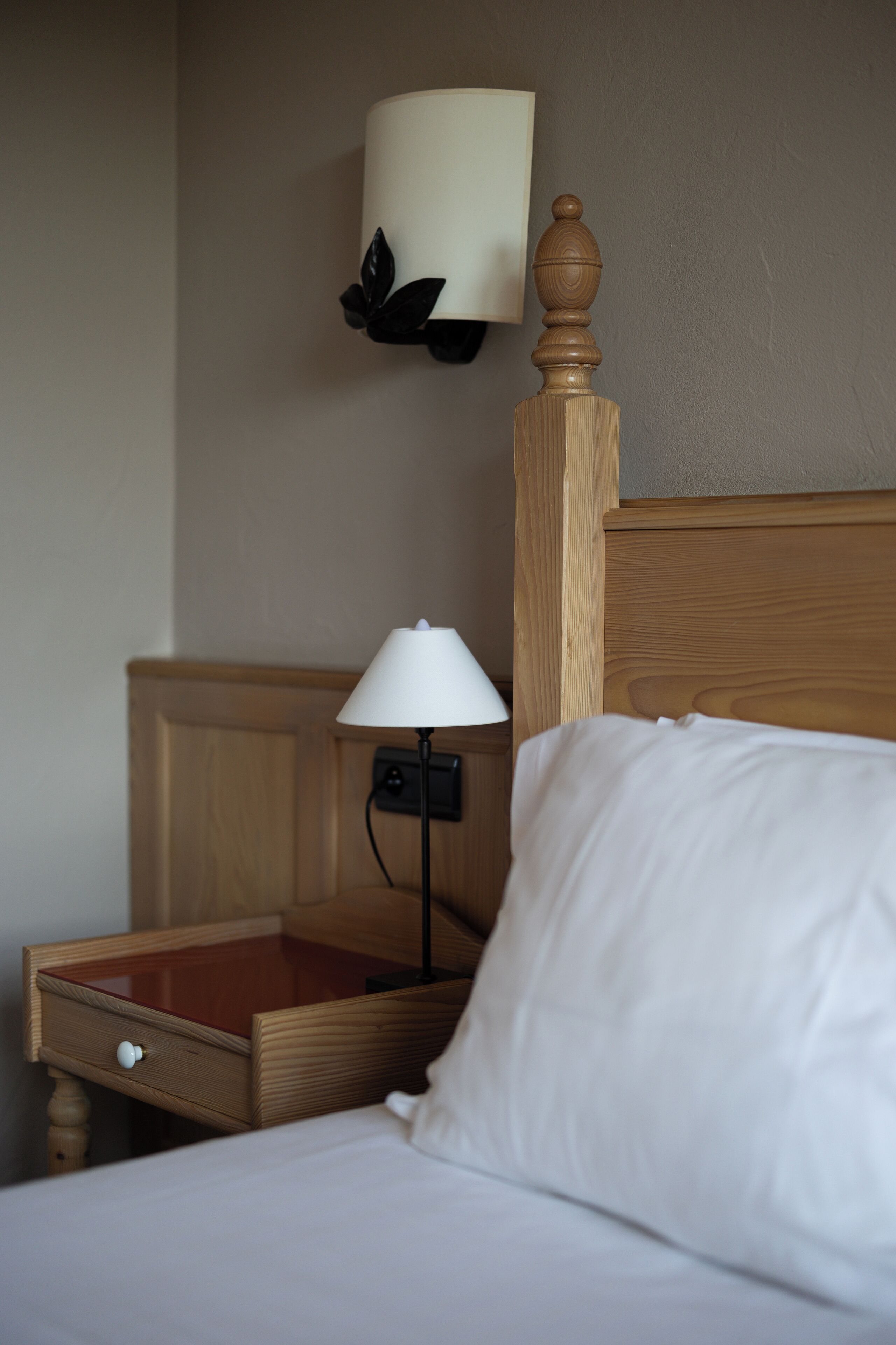 A neatly made bed with white linens, a wooden headboard, a bedside table with a lamp, and a wall-mounted light fixture.