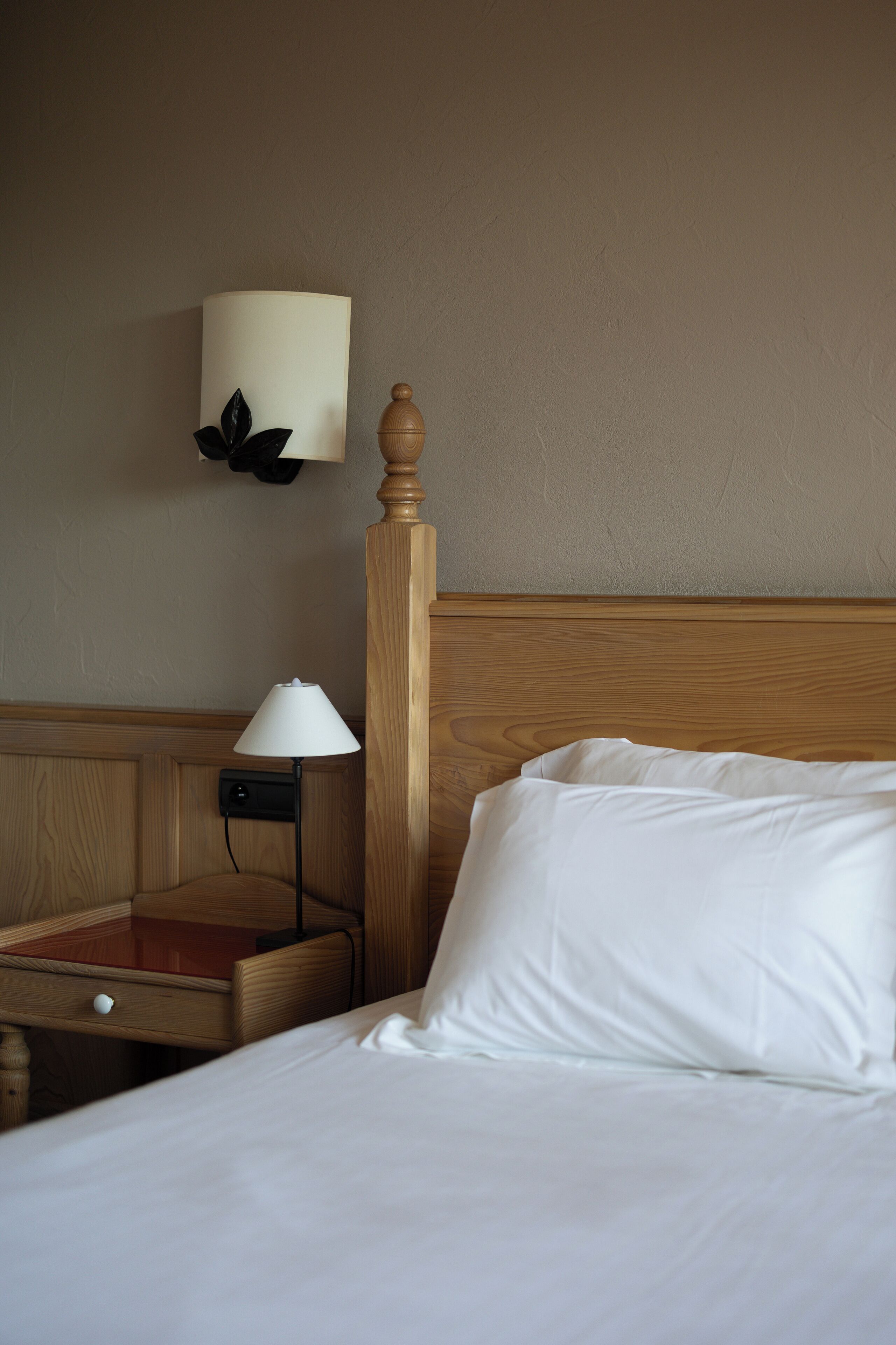 A neatly made bed with white linens, a wooden headboard, a bedside table with a lamp, and a wall-mounted light fixture.