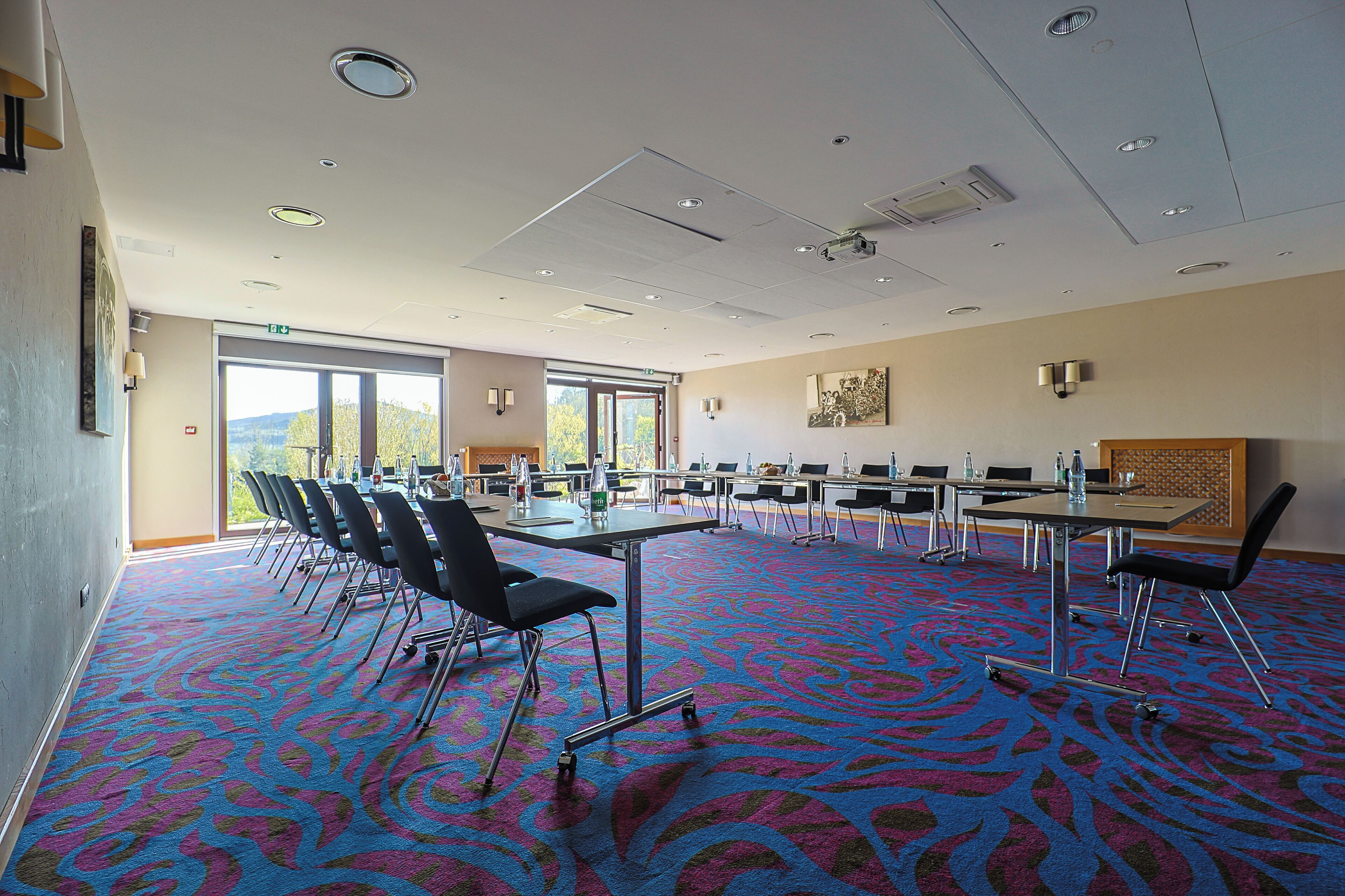 A conference room with a patterned carpet, multiple tables, chairs, and a large window offering a view of the outdoors.