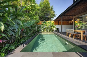 A rectangular swimming pool surrounded by lush tropical plants, with a wooden deck and outdoor seating area on one side.