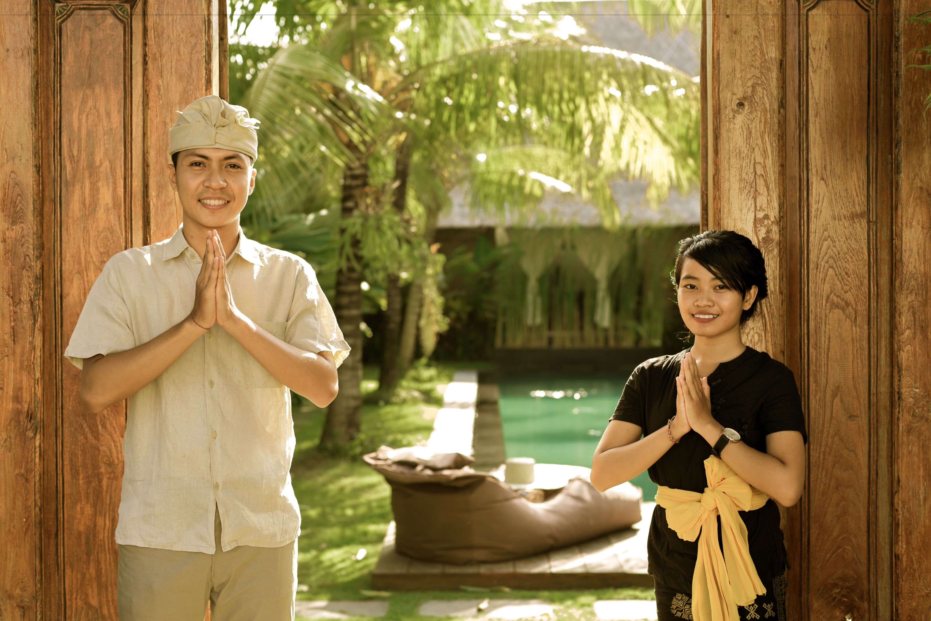 A man and a woman standing in a wooden doorway with a pool and palm trees in the background.