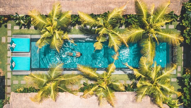 Vue aérienne d’une piscine entourée de palmiers, avec une personne qui nage et une chaise longue sur le côté gauche.