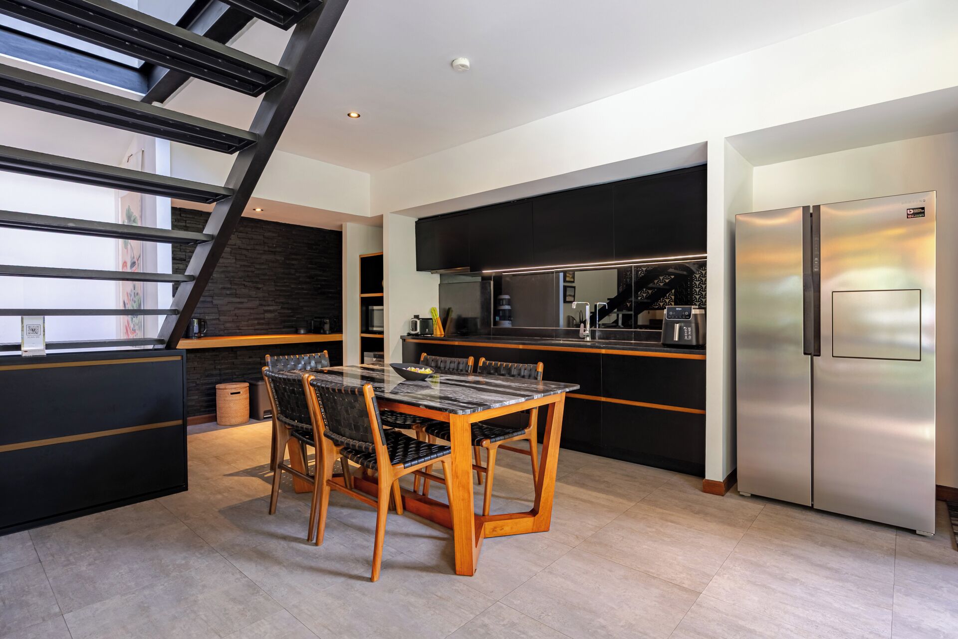 A modern kitchen with a built-in refrigerator, a dining area with a marble table and wooden chairs, and a staircase with a black railing.