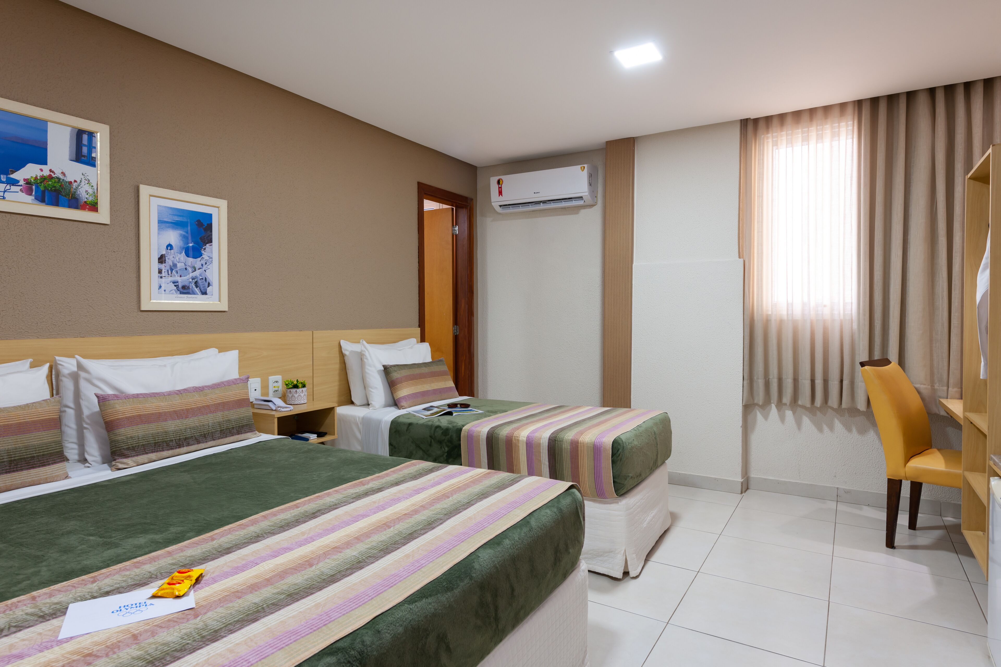 Une chambre d’hôtel avec deux lits, un bureau, une chaise, un climatiseur et deux tableaux encadrés au mur.